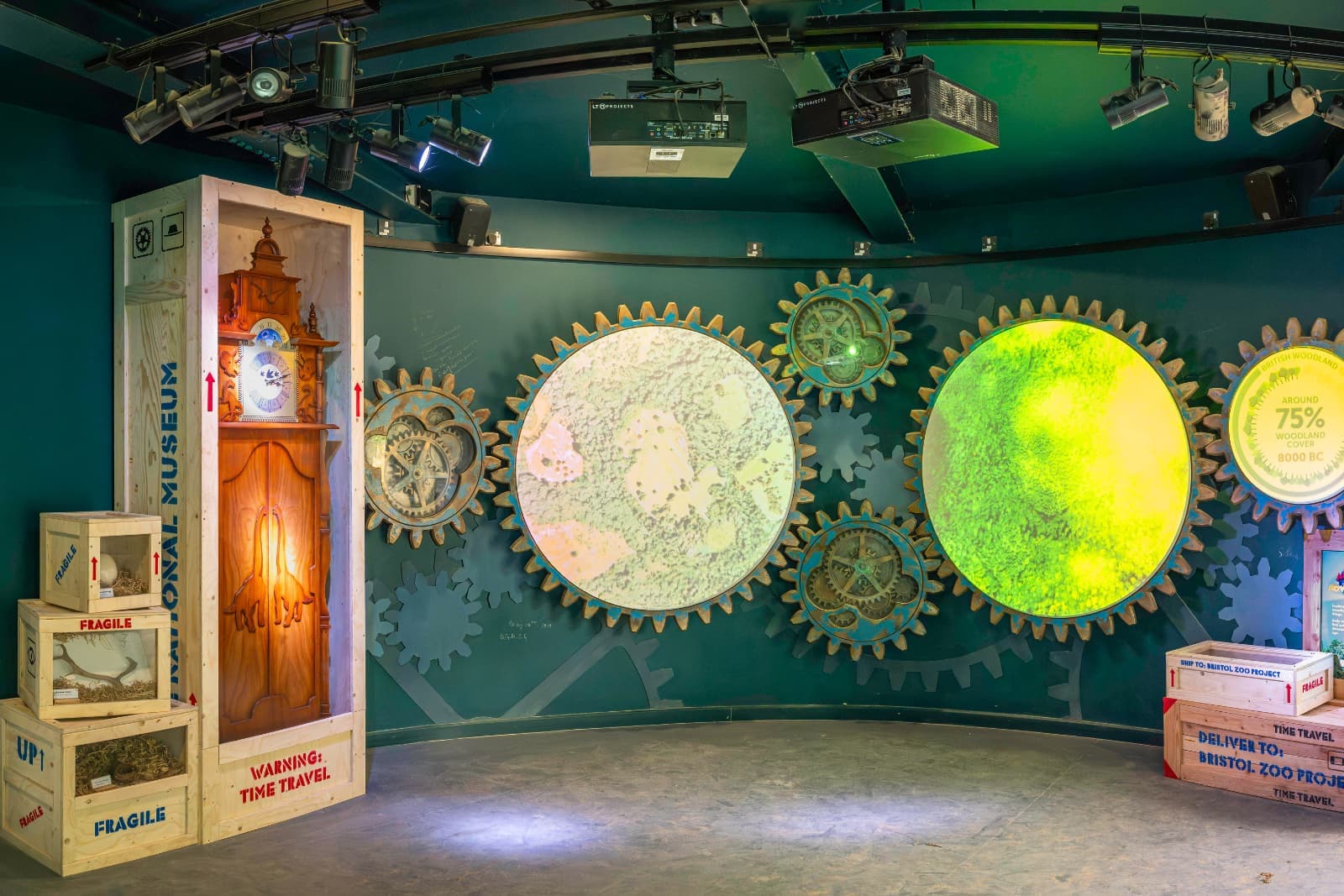Museum exhibit with large, illuminated gears and maps on a wall. Wooden crates labeled "fragile" and "time travel" are on the floor.
