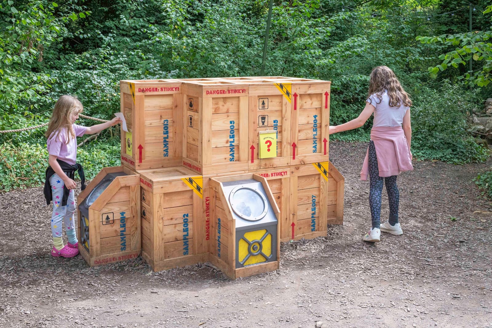 Two girls interact with stacked wooden crates labeled "Danger" and featuring symbols, in a wooded area.