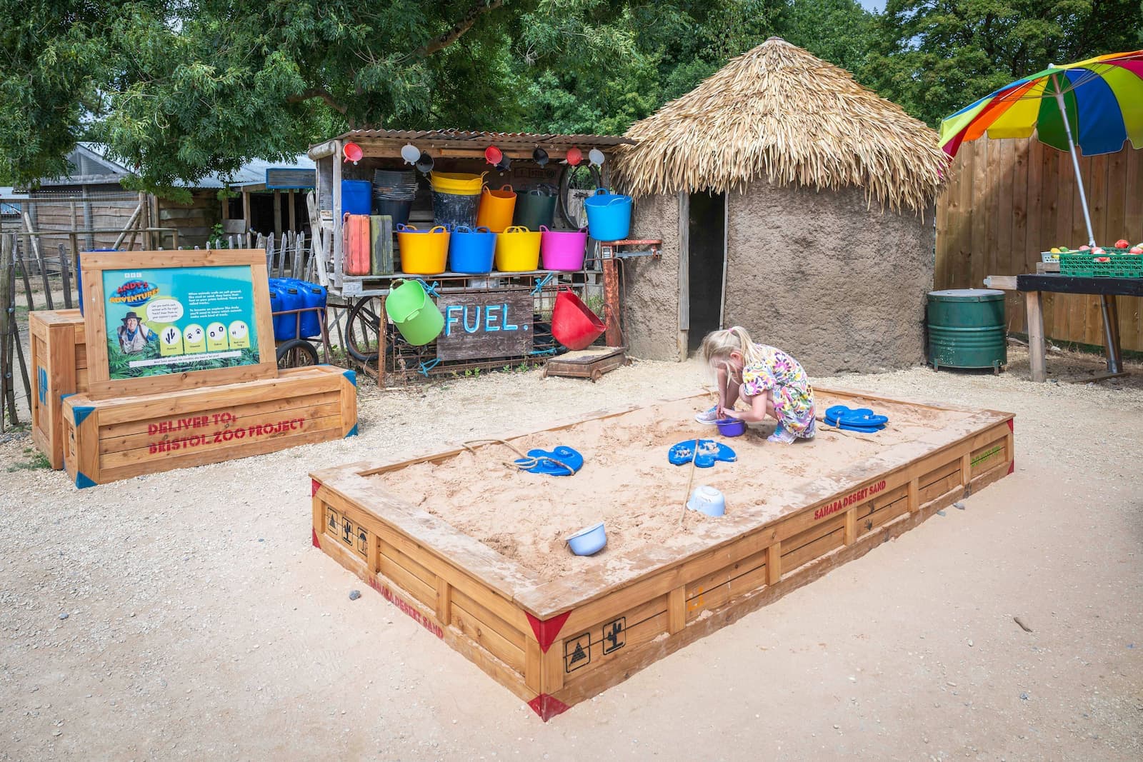 Child playing in a sandbox with toys, surrounded by colorful buckets, a thatched hut, and a signboard in an outdoor setting.
