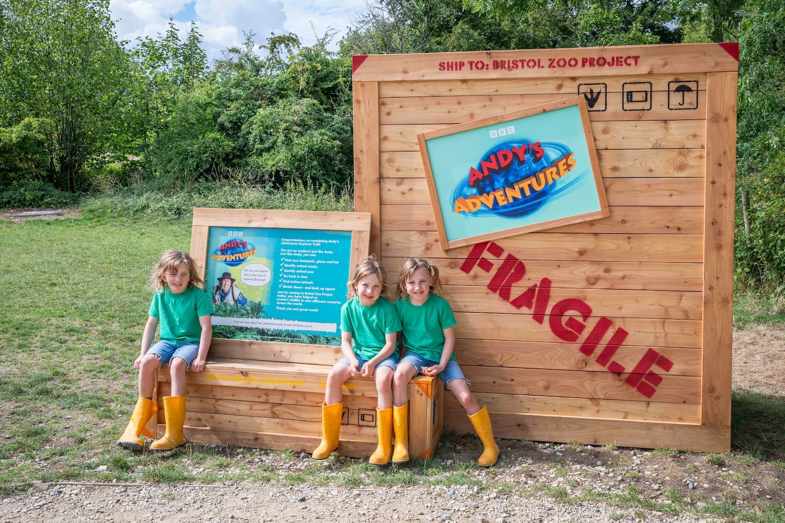 Three children in green shirts and yellow boots sit on a wooden bench next to a large "Andy's Adventures" sign outdoors.