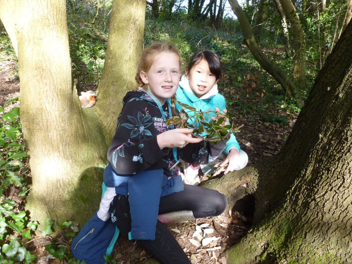 Two young girls sit crouched at the base of a tree, with one holding a wreath