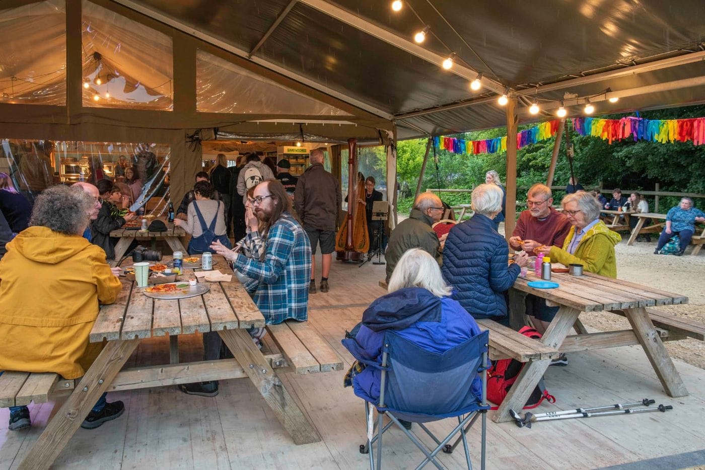 Groups of people sit on wooden benches in a covered outdoor space, eating pizza