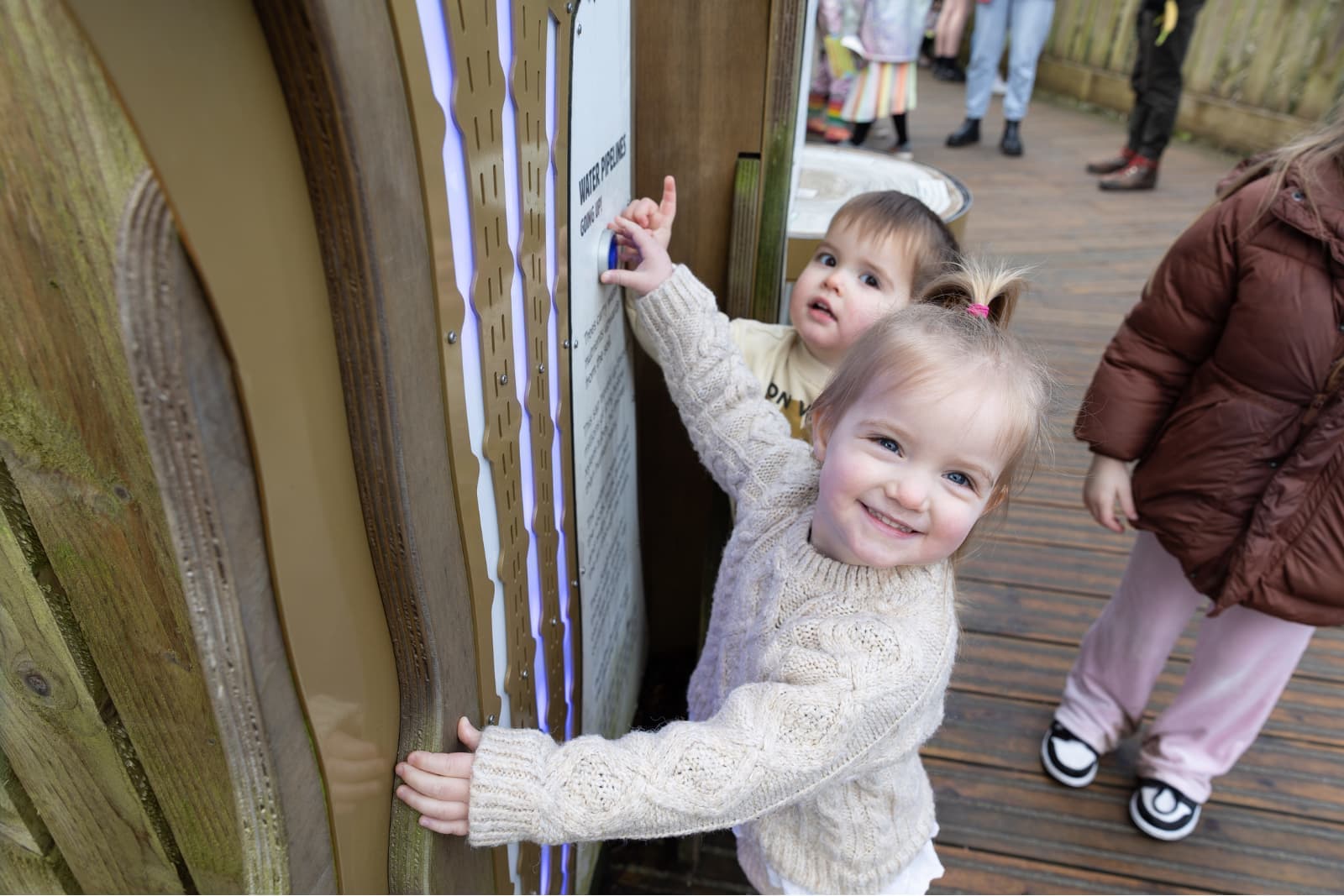 Smiling toddler in a knit sweater reaches a play panel while another child behind presses a button on a wooden deck.