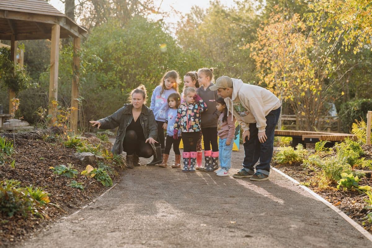 A group of children and two adults explore a garden path, with one adult pointing at something on the ground. Trees and a gazebo are in the background.